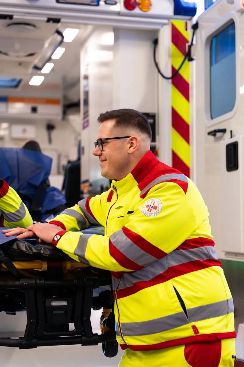 Ein Sanitäter steht vor einem Rettungswagen angelehnt an die ausgefahrene Trage, schaut nach links und lächelt.