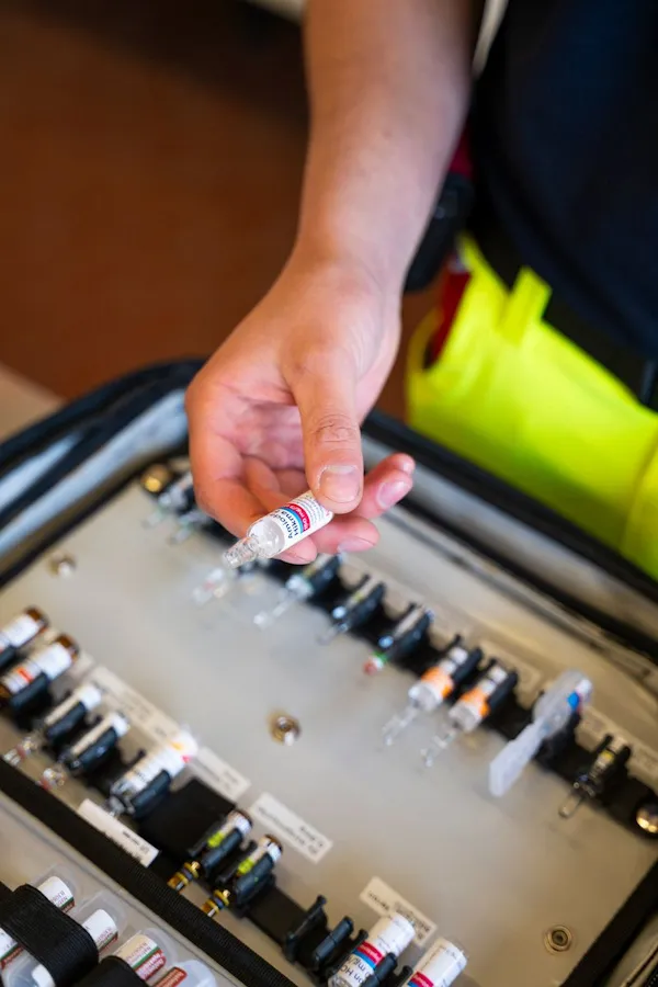 Nahaufnahme einer Hand, die eine Ampulle eines Medikaments hochhält. Im Hintergrund ist ein Ampullarium zu sehen mit weiteren Medikamenten