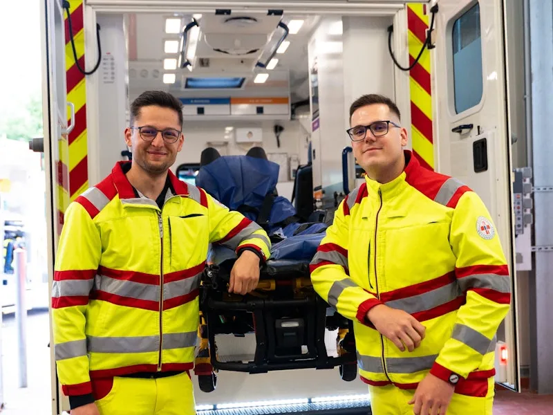 Zwei Sanitäter in gelb-roter Dienstkleidung stehen vor einem Rettungswagen mit offener Hecktür. Beide lehnen sich an die ausgefahrene Trage an und lächeln in die Kamera